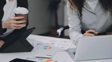 Beyaz ceketli ve gözlüklü genç bir kadın iş arkadaşıyla ofiste oturuyor ve elinde bir fincan kahve ve dizüstü bilgisayar kullanıyor..