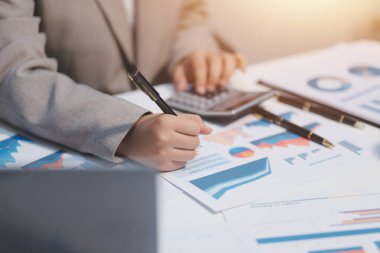 Hesap makinesi ve yazı kullanan bir iş kadını, hesap makinesiyle not alır. Ofiste çalışan bir kadın. Masasında dizüstü bilgisayar ve belgeler var.