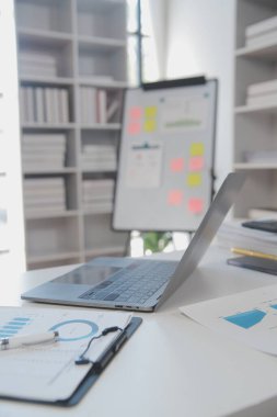 Close up view of simple workspace with laptop, notebooks, coffee cup and tree pot on white table with blurred office room background