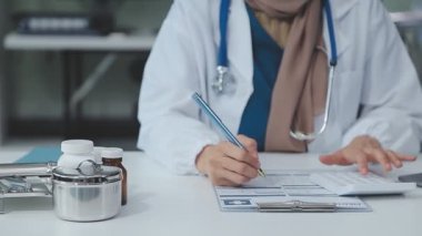 Genç bayan doktor notlar yazıyor ve ofisinde notlar alıyor. Masanın yanında oturuyor.