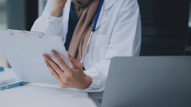 Doktor önlüklü masada oturuyor ve elinde bir pano var.
