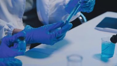 Koruyucu eldivenlerle laboratuvar asistanı elinde mavi sıvıyla tüpü tutuyor.