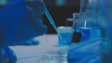 Kapat. Mavi eldivenli laboratuvar asistanı elinde sıvı bir test tüpü tutuyor.