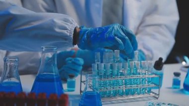 Laboratuvar asistanı elinde matara ve mavi sıvı test tüpleri tutuyor.