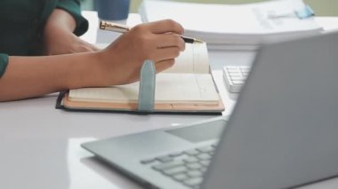 Yakın plan - dizüstü bilgisayar kullanan genç bir kadının, elinde defteriyle masada otururken görüntüsü