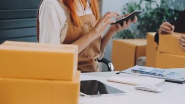 Dizüstü bilgisayarda çalışan Asyalı bir kadın ve elinde akıllı telefonlu karton bir paket kutusu var. dağıtım hizmeti, çevrimiçi alışveriş, iş konsepti.