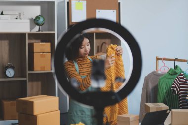 Vlogger, blog yazarı, küçük KOBİ iş kadını güzel bir kadın cep telefonuyla kıyafetleri satıyor canlı yayın satışı yapıyor, ana ofiste ürün sunumu detaylarını gösteriyor, girişimci ekommerce