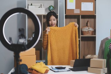 Vlogger, blog yazarı, küçük KOBİ iş kadını güzel bir kadın cep telefonuyla kıyafetleri satıyor canlı yayın satışı yapıyor, ana ofiste ürün sunumu detaylarını gösteriyor, girişimci ekommerce