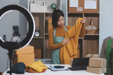 Vlogger, blog yazarı, küçük KOBİ iş kadını güzel bir kadın cep telefonuyla kıyafetleri satıyor canlı yayın satışı yapıyor, ana ofiste ürün sunumu detaylarını gösteriyor, girişimci ekommerce