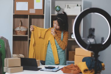 Vlogger, blog yazarı, küçük KOBİ iş kadını güzel bir kadın cep telefonuyla kıyafetleri satıyor canlı yayın satışı yapıyor, ana ofiste ürün sunumu detaylarını gösteriyor, girişimci ekommerce