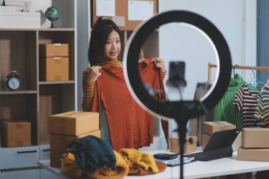 Vlogger, blog yazarı, küçük KOBİ iş kadını güzel bir kadın cep telefonuyla kıyafetleri satıyor canlı yayın satışı yapıyor, ana ofiste ürün sunumu detaylarını gösteriyor, girişimci ekommerce