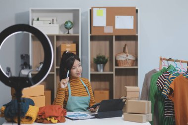 Vlogger, blog yazarı, küçük KOBİ iş kadını güzel bir kadın cep telefonuyla kıyafetleri satıyor canlı yayın satışı yapıyor, ana ofiste ürün sunumu detaylarını gösteriyor, girişimci ekommerce