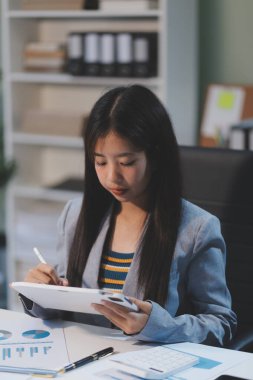 Güzel Asyalı iş kadını, ofise yerleştirilmiş evraklarla birlikte tablet ve dizüstü bilgisayar kullanıyor..