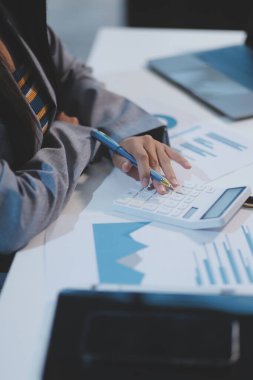 Ofiste oturan güzel Asyalı kadın, hesap makinesi, veriler, çizelgeler ve dokümanlar üzerinde çalışıyor iş yerinde masanın üzerinde. Kameraya bak.