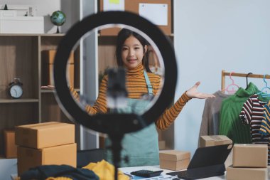 Vlogger, blog yazarı, küçük KOBİ iş kadını güzel bir kadın cep telefonuyla kıyafetleri satıyor canlı yayın satışı yapıyor, ana ofiste ürün sunumu detaylarını gösteriyor, girişimci ekommerce