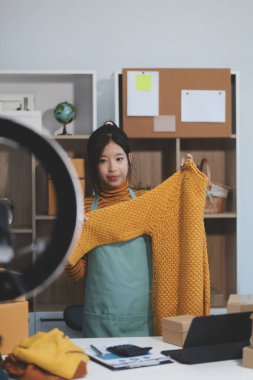 Vlogger, blog yazarı, küçük KOBİ iş kadını güzel bir kadın cep telefonuyla kıyafetleri satıyor canlı yayın satışı yapıyor, ana ofiste ürün sunumu detaylarını gösteriyor, girişimci ekommerce