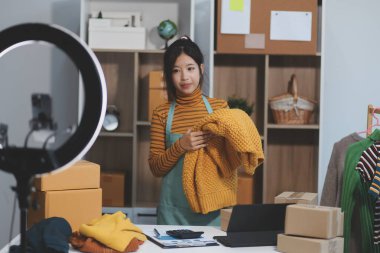 Vlogger, blog yazarı, küçük KOBİ iş kadını güzel bir kadın cep telefonuyla kıyafetleri satıyor canlı yayın satışı yapıyor, ana ofiste ürün sunumu detaylarını gösteriyor, girişimci ekommerce