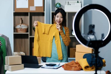 Vlogger, blog yazarı, küçük KOBİ iş kadını güzel bir kadın cep telefonuyla kıyafetleri satıyor canlı yayın satışı yapıyor, ana ofiste ürün sunumu detaylarını gösteriyor, girişimci ekommerce