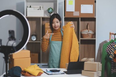 Vlogger, blog yazarı, küçük KOBİ iş kadını güzel bir kadın cep telefonuyla kıyafetleri satıyor canlı yayın satışı yapıyor, ana ofiste ürün sunumu detaylarını gösteriyor, girişimci ekommerce