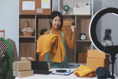 Vlogger, blog yazarı, küçük KOBİ iş kadını güzel bir kadın cep telefonuyla kıyafetleri satıyor canlı yayın satışı yapıyor, ana ofiste ürün sunumu detaylarını gösteriyor, girişimci ekommerce