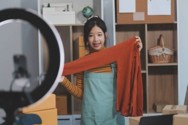 Vlogger, blog yazarı, küçük KOBİ iş kadını güzel bir kadın cep telefonuyla kıyafetleri satıyor canlı yayın satışı yapıyor, ana ofiste ürün sunumu detaylarını gösteriyor, girişimci ekommerce