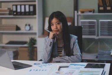 Ofiste oturan güzel Asyalı kadın, hesap makinesi, veriler, çizelgeler ve dokümanlar üzerinde çalışıyor iş yerinde masanın üzerinde. Kameraya bak.