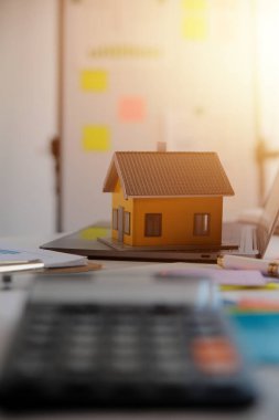 Diz üstü bilgisayarı olan ahşap masa, kalem, ev modeli ve yeşil ağaç hesap makinesi olan ev anahtarları, emlak konsepti. Resimleri kapat