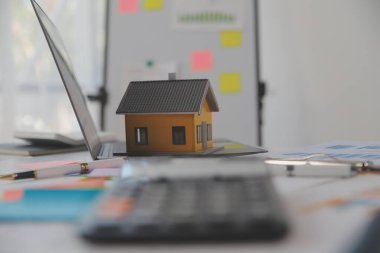 Diz üstü bilgisayarı olan ahşap masa, kalem, ev modeli ve yeşil ağaç hesap makinesi olan ev anahtarları, emlak konsepti. Resimleri kapat
