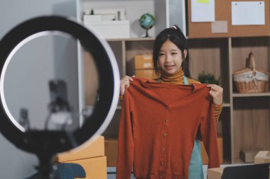 Vlogger, blog yazarı, küçük KOBİ iş kadını güzel bir kadın cep telefonuyla kıyafetleri satıyor canlı yayın satışı yapıyor, ana ofiste ürün sunumu detaylarını gösteriyor, girişimci ekommerce