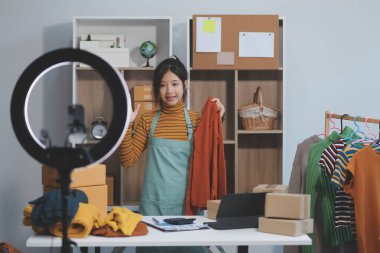 Vlogger, blog yazarı, küçük KOBİ iş kadını güzel bir kadın cep telefonuyla kıyafetleri satıyor canlı yayın satışı yapıyor, ana ofiste ürün sunumu detaylarını gösteriyor, girişimci ekommerce