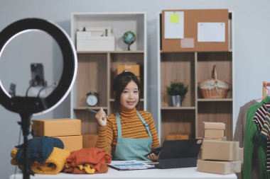 Vlogger, blog yazarı, küçük KOBİ iş kadını güzel bir kadın cep telefonuyla kıyafetleri satıyor canlı yayın satışı yapıyor, ana ofiste ürün sunumu detaylarını gösteriyor, girişimci ekommerce