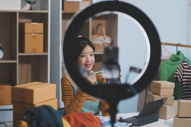 Vlogger, blog yazarı, küçük KOBİ iş kadını güzel bir kadın cep telefonuyla kıyafetleri satıyor canlı yayın satışı yapıyor, ana ofiste ürün sunumu detaylarını gösteriyor, girişimci ekommerce