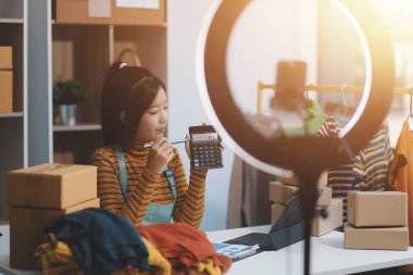 Vlogger, blog yazarı, küçük KOBİ iş kadını güzel bir kadın cep telefonuyla kıyafetleri satıyor canlı yayın satışı yapıyor, ana ofiste ürün sunumu detaylarını gösteriyor, girişimci ekommerce