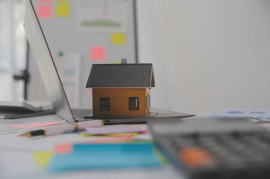 Diz üstü bilgisayarı olan ahşap masa, kalem, ev modeli ve yeşil ağaç hesap makinesi olan ev anahtarları, emlak konsepti. Resimleri kapat