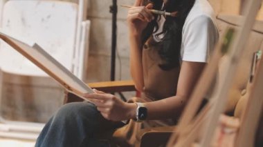 Beyaz gömlekli ve kotlu genç bir kadın ahşap masada elinde bir defterle oturuyor ve kalem çizimleri yapıyor..