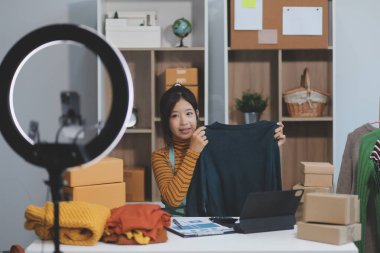 Vlogger, blog yazarı, küçük KOBİ iş kadını güzel bir kadın cep telefonuyla kıyafetleri satıyor canlı yayın satışı yapıyor, ana ofiste ürün sunumu detaylarını gösteriyor, girişimci ekommerce