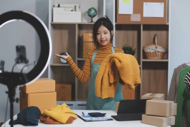 Vlogger, blog yazarı, küçük KOBİ iş kadını güzel bir kadın cep telefonuyla kıyafetleri satıyor canlı yayın satışı yapıyor, ana ofiste ürün sunumu detaylarını gösteriyor, girişimci ekommerce