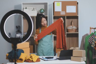 Vlogger, blog yazarı, küçük KOBİ iş kadını güzel bir kadın cep telefonuyla kıyafetleri satıyor canlı yayın satışı yapıyor, ana ofiste ürün sunumu detaylarını gösteriyor, girişimci ekommerce