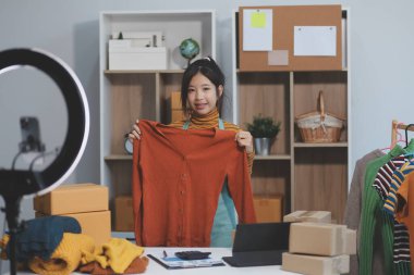 Vlogger, blog yazarı, küçük KOBİ iş kadını güzel bir kadın cep telefonuyla kıyafetleri satıyor canlı yayın satışı yapıyor, ana ofiste ürün sunumu detaylarını gösteriyor, girişimci ekommerce