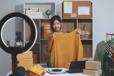 Vlogger, blog yazarı, küçük KOBİ iş kadını güzel bir kadın cep telefonuyla kıyafetleri satıyor canlı yayın satışı yapıyor, ana ofiste ürün sunumu detaylarını gösteriyor, girişimci ekommerce