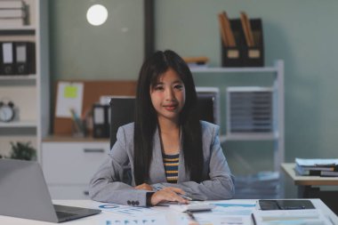 Ofiste oturan güzel Asyalı kadın, hesap makinesi, veriler, çizelgeler ve dokümanlar üzerinde çalışıyor iş yerinde masanın üzerinde. Kameraya bak.
