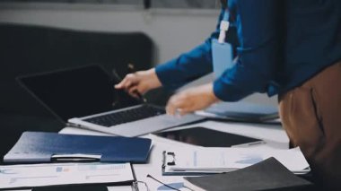 Takım elbiseli işadamı elinde belgelerle bilgisayarda çalışıyor.