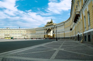 Genelkurmay binasının eğimli cephesinin yer aldığı Rusya 'nın Saint Petersburg kentindeki ikonik Saray Meydanı' nın geniş açılı bir görüntüsü. Karmaşık heykellerle süslenmiş kemer yolu parlak mavi bir gökyüzünün altında dağınık bulutlarla duruyor..