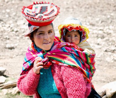 Renkli geleneksel el yapımı kıyafeti giymiş ve arkadan kovalayan bebeğini taşıyan gülümseyen bir Quechua kadın portresi kapatın. Ekim 21, 2012 - Patachancha, Cuzco, Peru