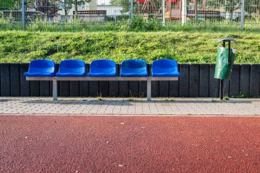 Boş mavi koltuklar, gün ışığında oyun parkının yanında yeşil çöp tenekesi olan bir bekleme alanı.
