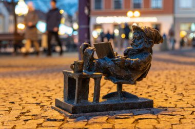 Wrocaw, Polonya - 01.18.2024: Bir bronz cüce heykeli, Wroclaw 's Market Square' de, küçük bir masanın yanındaki bir kitaba takılı olarak gelişigüzel duruyor. Canlı meydan yayalarla dolup taşıyor ve akşam atmosferinin tadını çıkarıyor..