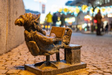 Wrocaw, poland - 01.18.2024: Büyüleyici bir bronz cüce Wroclaw 's Market Square' deki bir masada dizüstü bilgisayarla rahat bir şekilde oturuyor. İnsanlar yürürken meydan neşeli ışıklarla parıldıyor..
