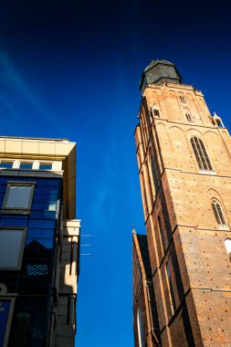 Tarihsel tuğla kule şehrin modern mimarisinin yanında berrak mavi gökyüzüne karşı dimdik ayakta duruyor.