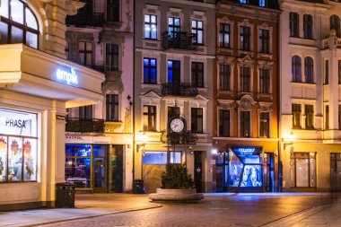 Ziyaretçiler, Torun 'un eski kasabasındaki Zegar saatinin etrafında toplanıyor. Canlı dükkanlar geceyi sıcak bir ışıltıyla aydınlatıyor..