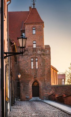 Çok iyi korunmuş bir tuğla kule, pastel gökyüzüne karşı yükselir, kaldırım taşları ve yakındaki binalara sıcak bir ışık saçar..